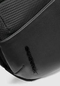 Zaino nero con texture e dettagli in pelle liscia, caratterizzato da un logo prominente e un compartimento con zip per una conservazione sicura.