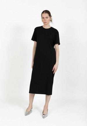 Frau in einem schwarzen, kurzärmeligen Midikleid und silbernen spitzen High Heels vor einem schlichten weißen Hintergrund stehend.