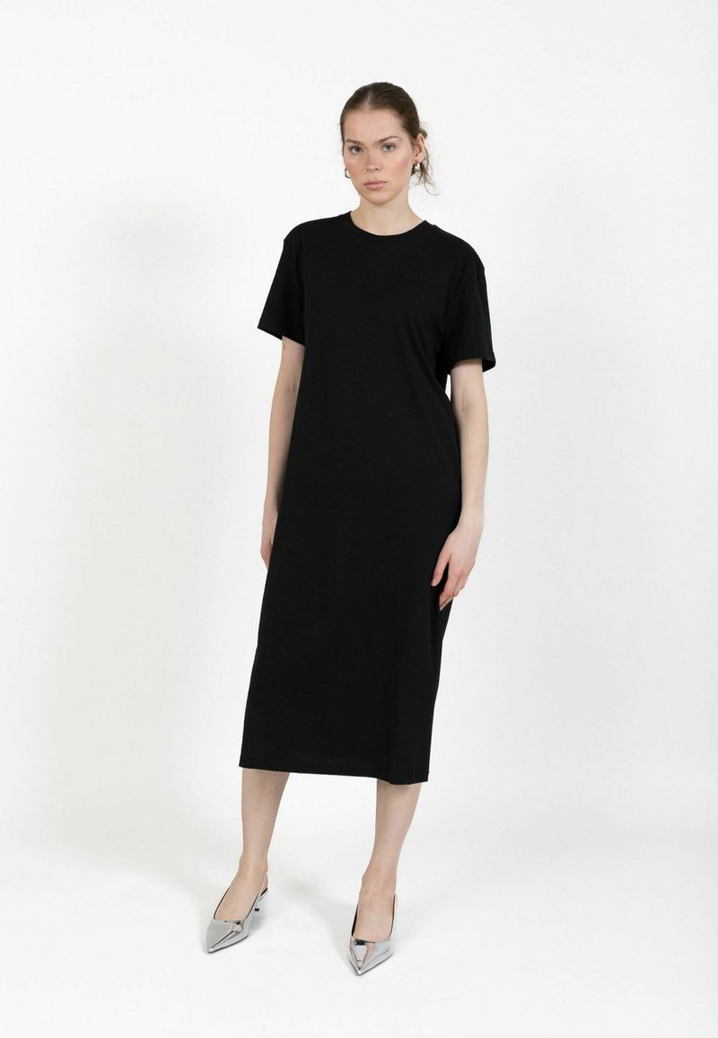 Kobieta stojąca na tle jednolitego białego tła, ubrana w czarną midi sukienkę z krótkim rękawem i srebrne szpilki o ostrym nosku.