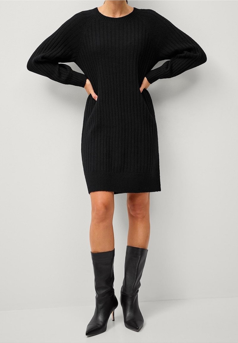 Robe pull noire en maille côtelée avec manches longues, encolure ronde et poches latérales, associée à des bottes noires à talons aiguilles montant jusqu'aux genoux.