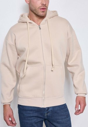 Homme portant un sweat à capuche beige zippé avec poches avant et cordons, associé à un jean bleu, debout devant un fond uni.