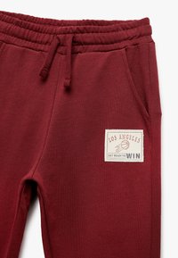 Burgunderfarbene Sweatpants mit elastischem Bund und Kordelzug. Mit einem weißen Stoffpatch, das "LOS ANGELES" und eine Basketballgrafik zeigt.