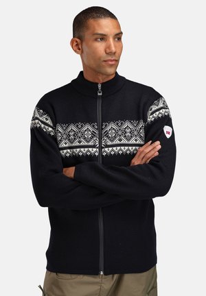 Mann med korslagte armer iført en svart genser med glidelås og hvite nordiske mønstre over brystet og ermene, med en lapp på venstre arm.