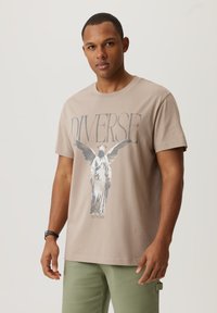 Beżowy t-shirt bawełniany z graficznym wzorem przedstawiającym posąg z skrzydłami oraz napisem "DIVERSE" powyżej. Krótkie rękawy i okrągły dekolt.