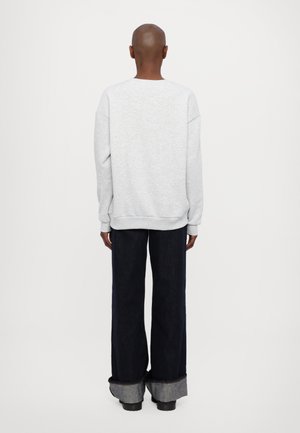 Person, der står med ryggen til iført en lysegrå sweatshirt og vidde mørke jeans med store manchetter over sorte sko.