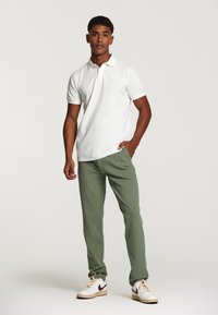 Cămașă polo albă, pantaloni verde măsliniu și adidași albi cu accente maronii. Design simplu, mâneci scurte, textură netedă. Fără modele vizibile.