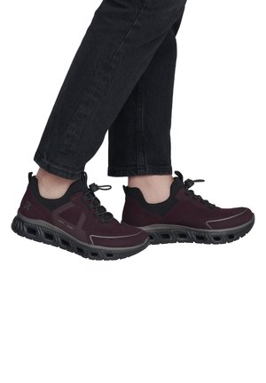Person trägt dunkelburgunderfarbene und schwarze Sportschuhe mit dicken Sohlen und schwarze Hose vor weißem Hintergrund.