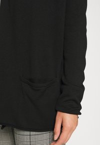 Zwarte longsleeve top gemaakt van gestructureerde stof, met een kleine zak aan de voorkant en een ontspannen pasvorm; gecombineerd met grijs geruite broek.