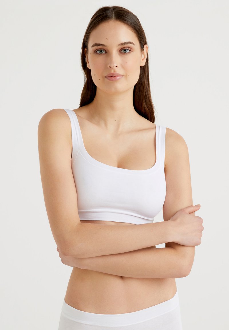 Bralette blanco de algodón con tirantes anchos y un diseño de escote redondo. Textura suave, forma ajustada y estilo minimalista. La modelo posa con los brazos cruzados.