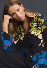 Blouse fleurie en tissu noir avec des couleurs vives, notamment vert, bleu et rouge, dotée d'un design à boutons et de manches longues.