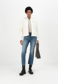 Gesteppte elfenbeinfarbene Bomberjacke mit gerippten Bündchen, kombiniert mit hochgeschnittenen blauen Jeans und schwarzen Schnürstiefeln, während sie eine dunkelgraue Handtasche hält.