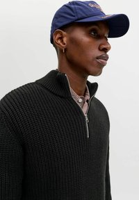 Junge Person mit einer marineblauen Mütze, kleinem Ringohrring, schwarzem strukturiertem Reißverschluss-Pullover über einem karierten Hemd, schaut nach rechts mit neutralem Ausdruck.