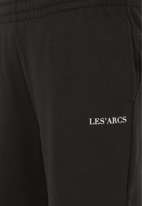 Jogging en coton noir avec une taille élastique, des poches latérales et un texte brodé blanc "LES'ARCS" sur la cuisse.