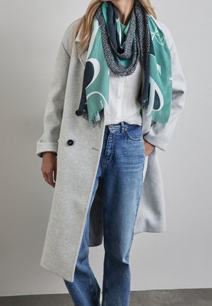 Femme portant un long manteau gris, une chemise blanche, un jean bleu et une écharpe à motifs bleu sarcelle drapée autour du cou.