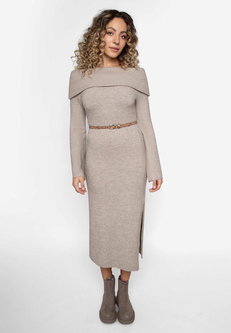 Stickad beige klänning med off-shoulder design, långa ärmar, ett bälte vid midjan och en sidospalt, som är ihop med grå ankelstövlar.