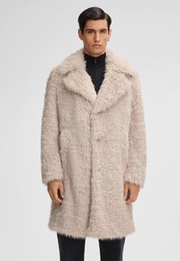 Uomo che indossa un cappotto in ecopelliccia beige chiaro con ampi risvolti sopra una maglietta nera con zip, in piedi di fronte a uno sfondo grigio uniforme.