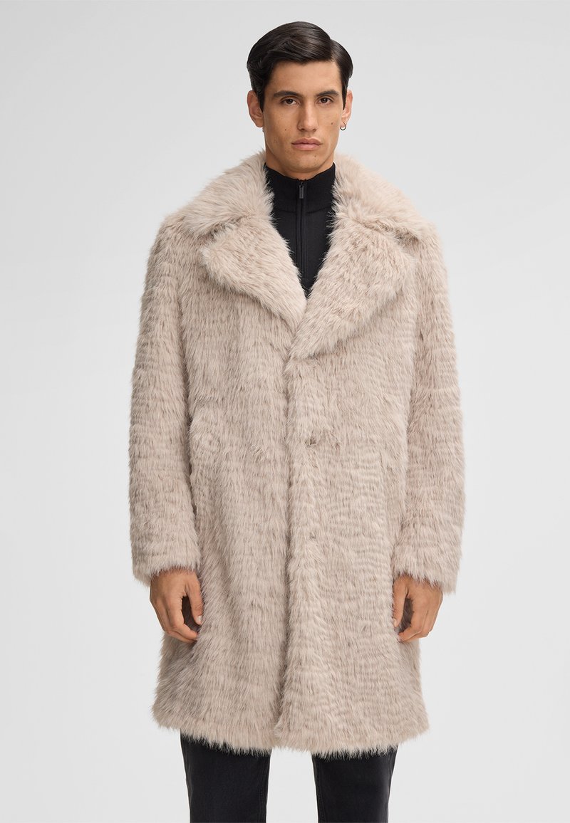 Uomo che indossa un cappotto in ecopelliccia beige chiaro con ampi risvolti sopra una maglietta nera con zip, in piedi di fronte a uno sfondo grigio uniforme.