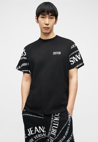 Νέος άνδρας που φοράει μαύρο μπλουζάκι και παντελόνι Versace Jeans Couture με λευκά μοτίβα λογότυπου, στέκεται με τα χέρια στις τσέπες.