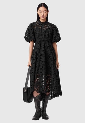 Kobieta w czarnej sukience midi z ażurowej koronki, z bufkami na rękawach, trzymająca czarną torebkę i nosząca czarne kowbojskie kozaki za kolano, stojąca.