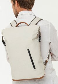 Zaino a rotolo di colore beige chiaro con un accento in pelle alla base, tasca con cerniera sul davanti, spallacci regolabili e un manico marrone.