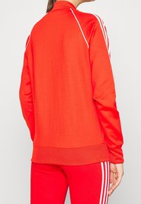 Červená sportovní bunda s vysokým límcem, dlouhými rukávy a bílými pruhy podél stran. Vyrobena z hladkého, elastického materiálu.