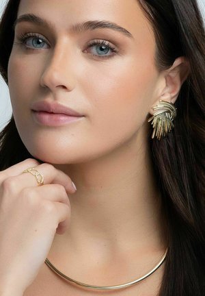 Guld hängehängen med ett lager på lager-design, ett slätt guldhalsband och en delikat ring med ädelstenar, alla bärs av en modell.