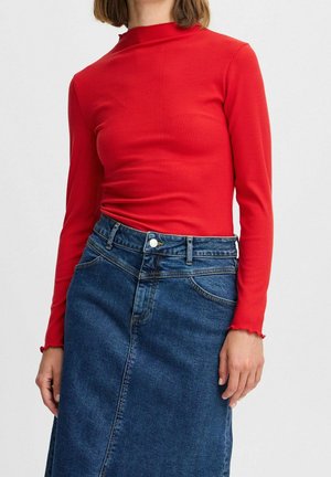 Frau trägt ein eng anliegendes, rotes, geripptes Langarmoberteil und einen hoch taillierten, dunkelblauen Jeansrock vor einfachem Hintergrund.