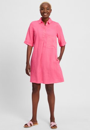 Roze linnen hemdjurk met een kraag, opgerolde mouwen, borstzak en zijzakken. Model draagt lichtroze slide sandalen.