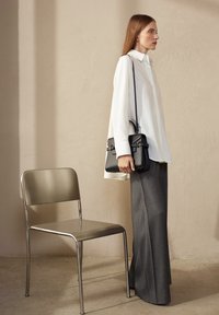 Borsa a spalla nera con design strutturato e doppie cinghie, camicia bianca a maniche lunghe e pantaloni grigi a gamba larga accanto a una sedia in metallo argentato.