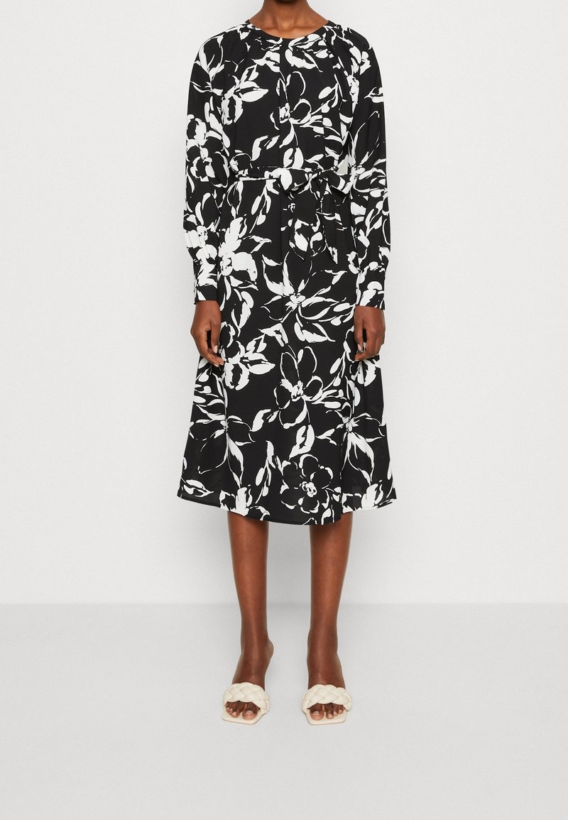 Robe noire longueur genoux avec un grand imprimé floral blanc, manches longues et ceinture nouée à la taille, portée avec des sandales blanches tressées à bout ouvert.
