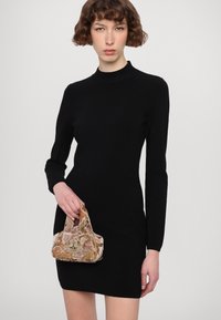 Svart långärmad klänning med hög hals. Modellen håller en liten, texturerad handväska i dämpade färger med blommönster. Enkel, figurnära silhuett.