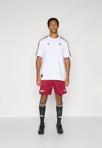 Witte T-shirt met korte mouwen en rode strepen op de schouders, gecombineerd met rode shorts met een logo. Zwarte sokken met witte strepen en zwarte schoenen.