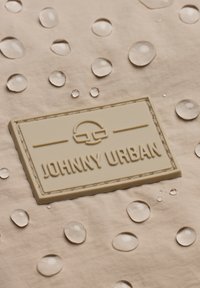 Tessuto beige con gocce d'acqua. Un'etichetta in gomma testurizzata recita "JOHNNY URBAN", con lettering in rilievo e un contorno liscio.