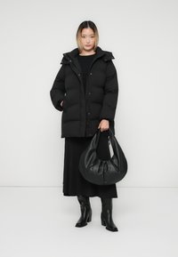 Frau in einer schwarzen Daunenjacke, schwarzem Kleid und schwarzen Stiefeln, die eine große schwarze Ledertasche vor einem schlichten weißen Hintergrund hält.