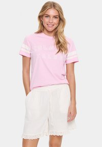 Helles rosa T-Shirt mit weißem "ISLAND TALES"-Text und gestreiften Ärmeln. Kombiniert mit weißen Shorts, die mit einem gewellten Ausschneidungsdetail am Saum versehen sind.