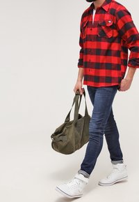 Camisa de franela a cuadros roja y negra, vaqueros azules, zapatillas deportivas blancas de caña alta y una bolsa de lona verde oliva con letras oscuras.