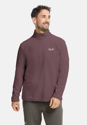 Man wearing a zipped maroon fleece pullover with Jack Wolfskin logo, standing and looking to the side against a plain background.
