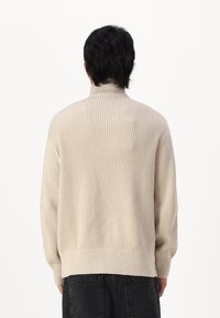 Kremno obarvan rebrast pulover s turtleneck izrezom, sproščenega kroja, z dolgimi rokavi in obrobenim robom, ki prikazuje teksturirani vzorec.