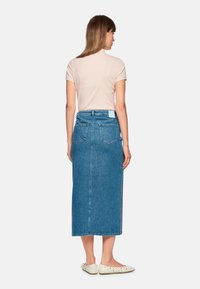 Denim maxi skirt in medium blue, featuring a straight cut, two back pockets, and a clean waistband. Paired with a light pink top and flat shoes.