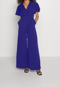 Mono vibrante azul con escote en V, mangas cortas y ondulantes, piernas anchas y bolsillos laterales. Tejido suave con un ajuste relajado y un detalle mínimo.