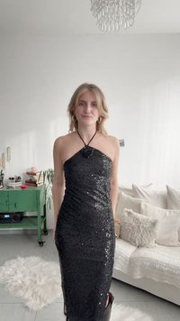 Γυναίκα με ξανθά μαλλιά που φοράει μαύρο φόρεμα με παγιέτες και ανοιχτή πλάτη, στέκεται σε ένα φωτεινό σαλόνι με λευκό καναπέ και πράσινο ντουλάπι.