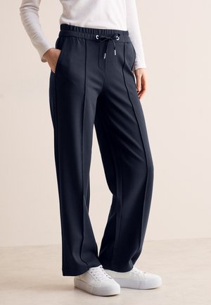 Pantalons de sport bleu marine avec une taille à cordon, poches latérales, détails de coutures verticales et une coupe décontractée, associés à des baskets blanches.