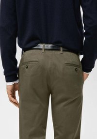 Pantaloni verde oliva con una texture liscia, caratterizzati da tasche posteriori e dettagli con bottoni, indossati con una maglia a maniche lunghe blu navy.