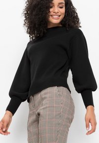Maglione lavorato a maglia nero con colletto alto e maniche a sbuffo. Abbinato a pantaloni a quadri con accenti rossi, che mostrano un tessuto testurizzato.
