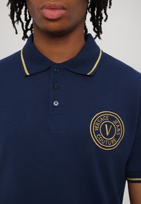 Marineblå polo med gule detaljer. Har krage, treknaps lukning og brodert logo på venstre bryst. Teksturert stoff.