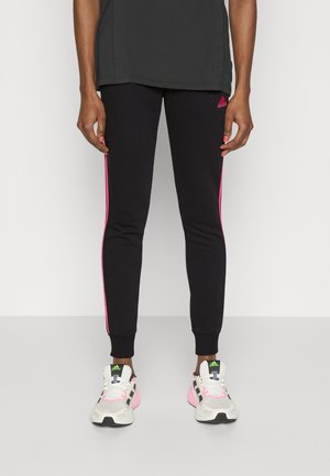 Persoon draagt zwarte Adidas legging met roze strepen en witte Adidas sneakers met roze accenten, staand tegen een effen achtergrond.