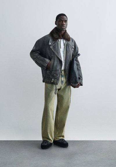 Sort læder motorcykeljakke med pelskrave, hvid skjorte, grønne bukser med vide ben og sorte broderede sko, stående mod en neutral baggrund.