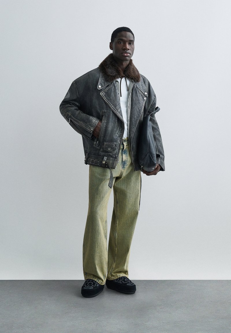Black leather motorcycle jacket with fur collar, white shirt, green wide-leg pants, and black embroidered shoes, standing against a neutral background.