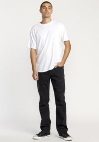 T-shirt blanc à manches courtes associé à un jean noir à jambes droites et des baskets noires, posé contre un fond clair uni.