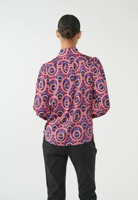Paarse blouse met een rood en roze cirkelpatroon, gestructureerde schouders en knopen. Gepaard met zwarte broeken, die een textuurrijke stof tonen.